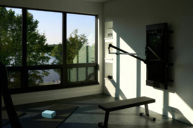 A bright, minimalist home gym with large black-framed windows overlooking a lake and trees. The room contains a wall-mounted strength-training machine, a flat workout bench, exercise mats, and small weights. Strong afternoon sunlight casts geometric shadows across the pale walls and grey floor.