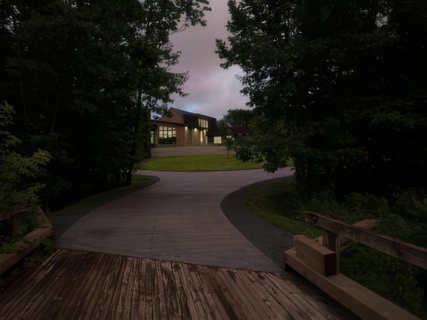 A dusk exterior view from a timber deck, looking along a curved paved driveway or walkway toward a contemporary house partially framed by dense mature trees. The house has a low-pitched roof, extensive glazing, and warm interior lighting. Timber decking and rustic timber railings occupy the foreground, with landscaped lawn and planting around the approach.