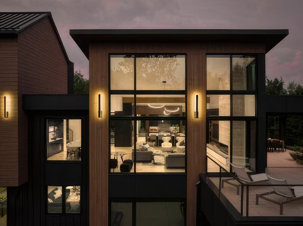 Twilight exterior view of a contemporary multi-level residence with dark vertical timber cladding, black-framed floor-to-ceiling glazing, and a projecting flat roof. The large windows reveal a warmly lit living area, while an elevated deck with outdoor lounge furniture occupies the right side. A connected side volume with matching dark finishes is visible on the left.