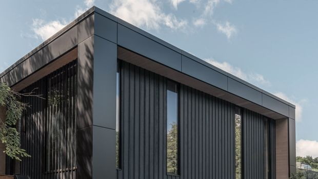 Exterior view of a contemporary single-storey building with a strong rectangular flat-roof form. The facade combines dark grey panel cladding with deep vertical ribbed screening, narrow vertical windows, and a recessed timber-toned soffit or trim. The composition is crisp and minimalist, with strong geometric lines and substantial framing around the openings.