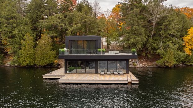 A contemporary two-level dark-clad waterfront house or boathouse set directly over a calm lake, surrounded by dense forest in autumn. The building has extensive floor-to-ceiling glazing, a flat roof, a broad upper terrace, and a timber deck extending over the water.