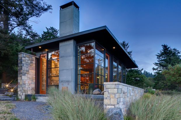 A modern single-storey pavilion-like house set among trees and grasses at dusk. The building has a broad flat roof with deep eaves, extensive floor-to-ceiling glazing, a tall exposed concrete chimney, natural stone walls and low stone balustrades, with warm interior lighting visible through the glass.