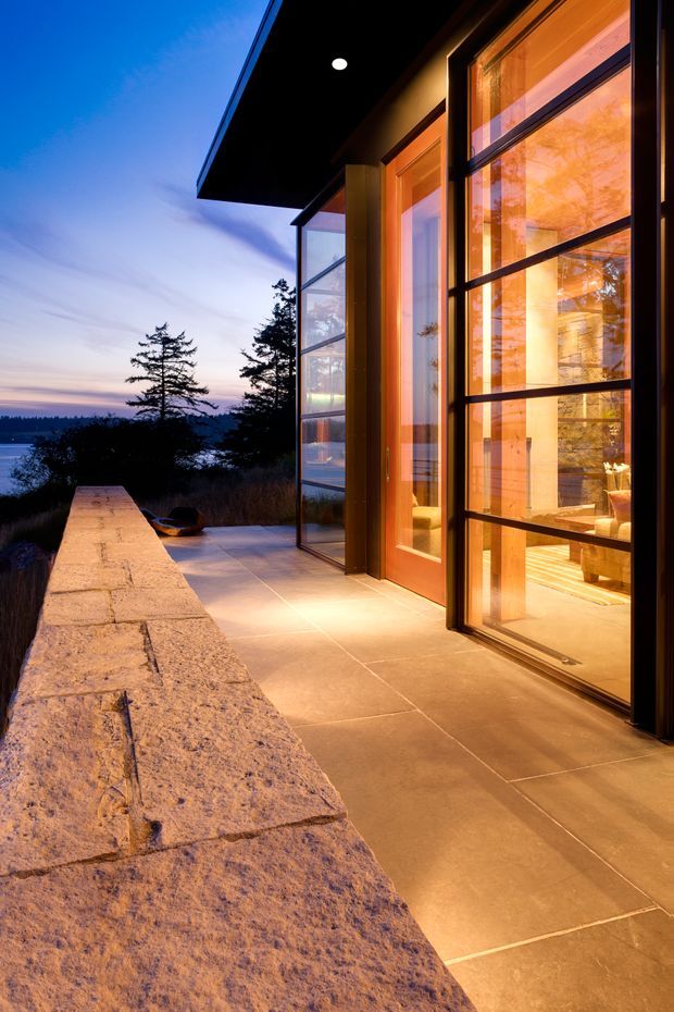 Twilight exterior view of a modern home opening onto a broad tiled terrace. Large black-framed glazed doors and windows reveal a warmly lit interior, while a substantial rough stone edge wall runs along the terrace. The home sits against a wooded waterfront or coastal-looking landscape, with strong warm interior lighting contrasting against the blue evening sky.