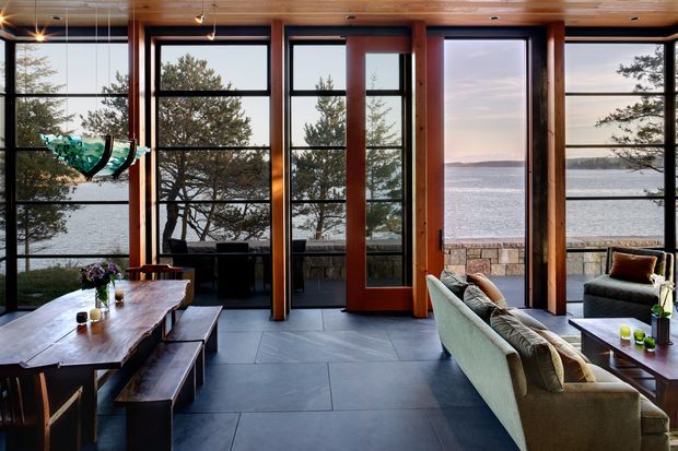 A warm, timber-lined living and dining room with expansive black-framed floor-to-ceiling glazing overlooking a calm waterfront. The room features a large solid timber dining table with bench seating, a neutral sofa and armchairs, dark large-format floor tiles, exposed timber posts and beams, and a sculptural pendant light. A stone terrace or retaining wall is visible beyond the glazing.