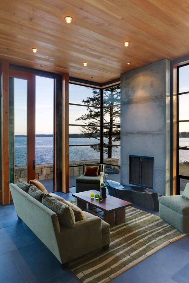 A refined living room with expansive floor-to-ceiling glazing overlooking a lake, furnished with pale upholstered sofas, a dark coffee table, and a striped area rug. The room features a warm timber-clad ceiling, exposed timber window and door frames, a large raw concrete fireplace, and dark tiled flooring. A balcony or terrace and waterfront landscape are visible beyond the glazing.