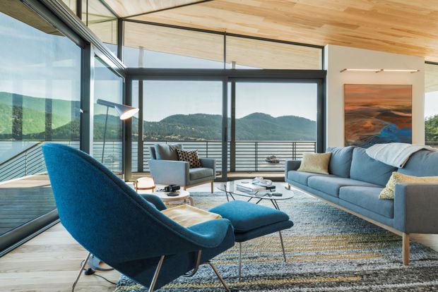 Bright, contemporary living room with floor-to-ceiling glazing opening onto a balcony or terrace and views of wooded hills. The room has a warm timber-lined ceiling and pale timber flooring, dark-framed sliding glass doors, a large blue-grey sofa, matching sculptural lounge chair and ottoman, patterned area rug, coffee tables, and modern wall art. The space feels airy, relaxed, and connected to the landscape.
