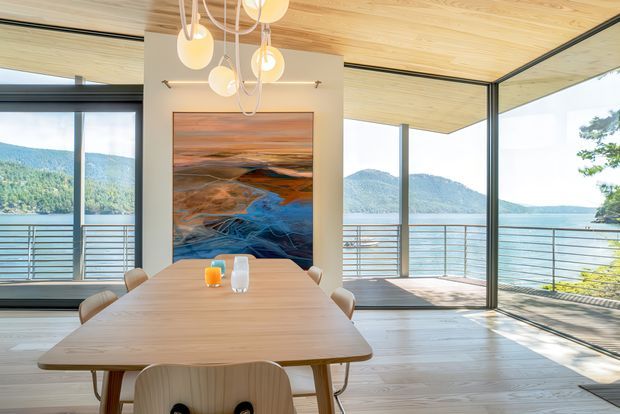 A bright contemporary dining area with a large light timber dining table and sculptural pendant lights. Full-height black-framed glazing opens to a balcony with metal railings and expansive views across a lake and wooded hills. The ceiling is clad in light timber, and a colourful landscape artwork forms a feature on the central wall.
