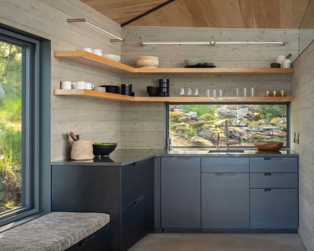 Compact, carefully designed kitchen or kitchenette with dark matte built-in cabinetry, a dark benchtop, open timber shelves, and pale timber wall and ceiling lining. Large windows frame a rocky landscaped garden, while integrated linear lighting and a built-in window seat add functionality. The space has a warm, contemporary cabin-like character with a restrained natural palette.