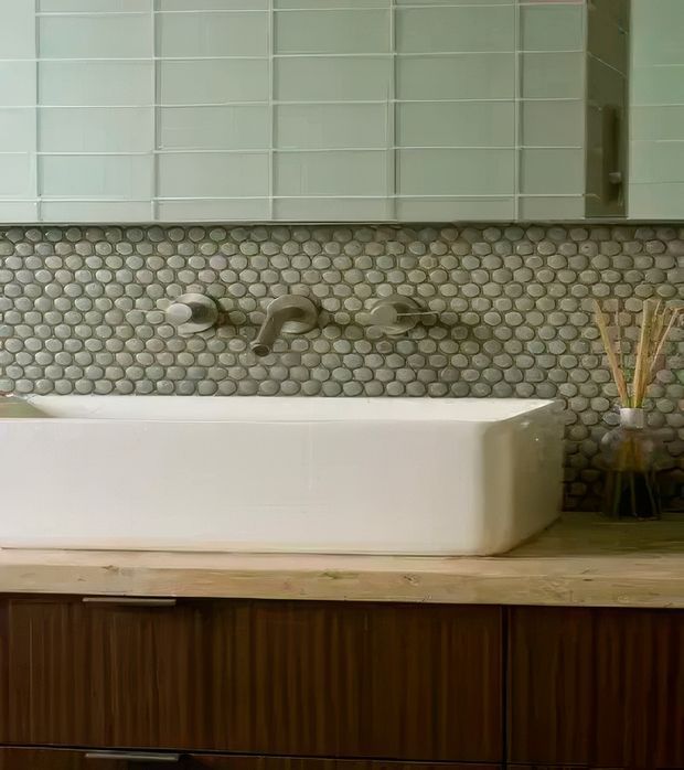 Close-up view of a refined bathroom featuring a freestanding white bathtub set into a timber vanity or cabinet base. The wall behind the tub has a rounded pebble mosaic tile backsplash with brushed metal bath fittings, while white rectangular wall tiles and mirrored cabinetry appear above. A small vase with decorative reeds sits at the side, adding a natural accent.