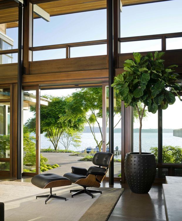 A double-height, lakefront living space with extensive dark-framed glazing and warm timber ceiling and wall detailing. The room contains a black leather lounge chair and ottoman, a large potted indoor tree, and a contemporary rug over a pale floor. Open doors connect to a landscaped terrace and water views beyond.