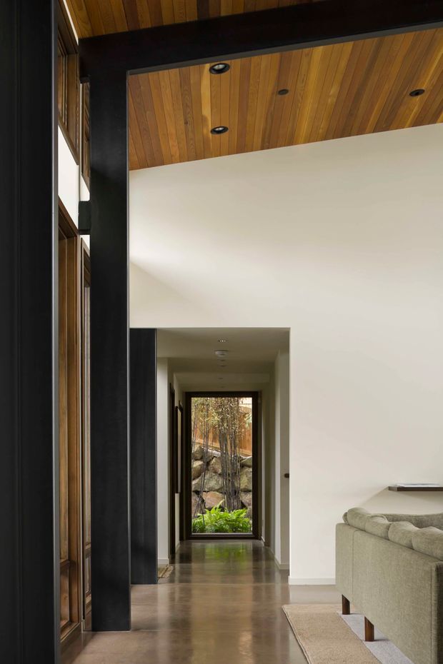 Interior view of a refined contemporary living area opening onto a long hallway. The space features a warm timber-lined ceiling with recessed downlights, exposed dark structural beams and trim, white plaster walls, polished dark timber flooring, and a large glazed opening at the end framing a planted outdoor area. A neutral upholstered sofa and rug are partially visible in the foreground.