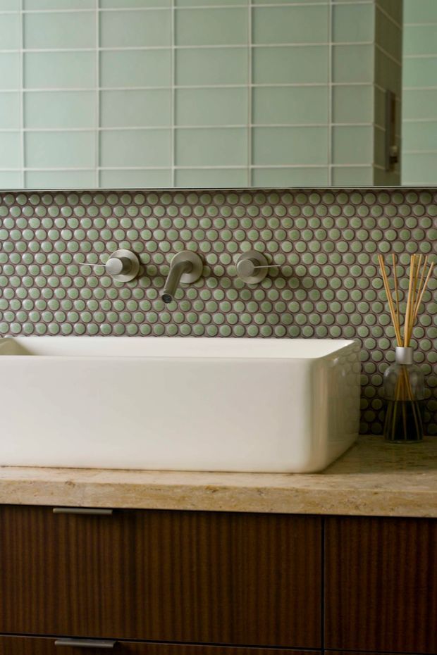 Close-up view of a refined bathroom vanity featuring a white vessel basin on a beige stone benchtop, dark ribbed timber cabinetry, brushed metal wall-mounted tapware, and a distinctive green circular mosaic tile splashback beneath pale rectangular wall tiles. A small decorative diffuser sits at the side.