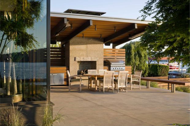 A contemporary outdoor entertaining area with a broad timber-lined roof canopy, exposed dark structural beams, a built-in masonry fireplace, outdoor dining furniture, barbecue cabinetry, and a long timber dining table. The space opens onto a paved terrace and landscaped suburban surroundings, with floor-to-ceiling glazing visible at the left.