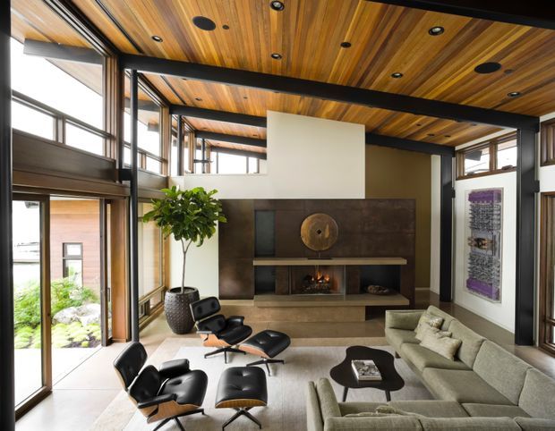 A spacious, refined living room with a large sectional sofa, sculptural lounge chairs, dark coffee table, and a substantial built-in fireplace set against a dark stone or textured feature wall. The room features extensive floor-to-ceiling glazing with dark frames, exposed black structural beams, and a warm timber-lined ceiling with recessed lighting and speakers. A potted indoor tree and artwork add softness and character.
