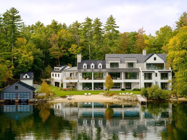 A large multi-storey residential house viewed across a calm lake, surrounded by dense mature woodland in autumn. The home has a light-coloured exterior, dark grey pitched and gabled roofs, extensive glazing, multiple balconies or terraces, and a separate dark garage or boathouse near the water. The lake and building are reflected in the foreground.