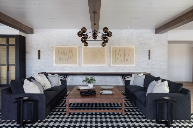 A spacious, refined living room with two dark charcoal sofas facing a substantial timber coffee table. The room features a bold black-and-white geometric rug, a white-painted brick feature wall with three framed artworks, dark timber ceiling beams, a long slim console, and a sculptural bronze multi-globe pendant light. A dark-framed glazed door or screen is visible at left, with an adjacent opening to another area at right.