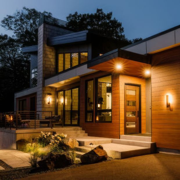 Twilight exterior view of a modern multi-level home with a strongly layered, angular roofline and projecting canopy. The facade combines warm horizontal timber cladding, dark metal trim, and pale masonry or concrete-block walls, with extensive amber-lit glazing and a timber entry door. Steps, paved terraces, low planting, and integrated exterior wall lights lead to the entrance, creating a warm and dramatic evening presentation.