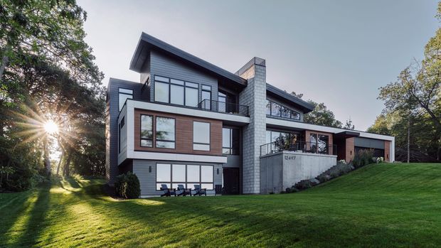 Large multi-level contemporary residence set within a landscaped, sloping lawn. The building features strongly projecting flat roof planes, dark horizontal cladding, extensive black-framed glazing, warm timber-toned wall panels, and a prominent pale masonry chimney or feature wall. The view is an exterior architectural photograph with mature trees and a broad lawn surrounding the house.