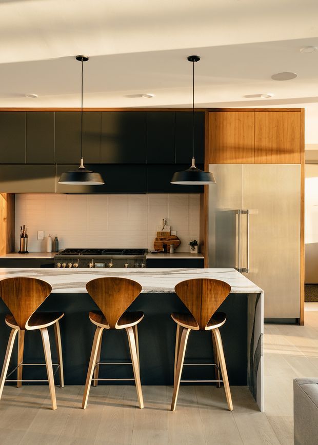 A contemporary kitchen viewed straight on, centred on a dark island with a pale stone benchtop and three sculptural timber bar stools. Matte dark cabinetry spans the rear wall, paired with warm timber cabinetry around a large stainless-steel refrigerator. Two black pendant lights hang above the island, while a pale tiled splashback, integrated cooktop and warm timber detailing complete the refined, high-contrast scheme.