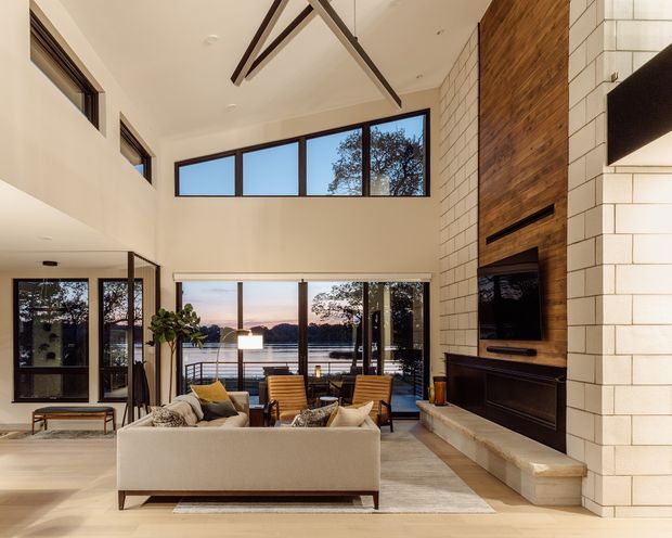A spacious contemporary living room with a double-height ceiling and expansive glazing overlooking a waterfront setting. The room features a large light upholstered sofa, timber lounge chairs, a sculptural suspended ceiling light, a white masonry fireplace wall with integrated dark cabinetry and television, and a warm timber fireplace surround. Exposed dark structural beams and high clerestory windows add architectural character.
