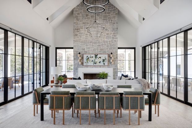 A bright, spacious open-plan dining and living area with a long dark dining table surrounded by upholstered chairs, positioned in front of a central natural-stone fireplace wall. The room has a high vaulted white ceiling with exposed beams, a series of circular pendant lights, extensive black-framed glazed doors and windows on both sides, pale flooring, and contemporary furnishings in neutral tones.