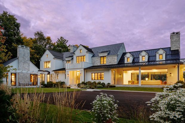 A large, multi-wing residential house photographed from the landscaped front garden at dusk. The home has pale stone and light painted exterior walls, dark pitched roofs with multiple dormer windows, several chimneys, warm illuminated windows, and a covered ground-floor veranda or entry zone. Mature planting and ornamental grasses surround the driveway and facade.