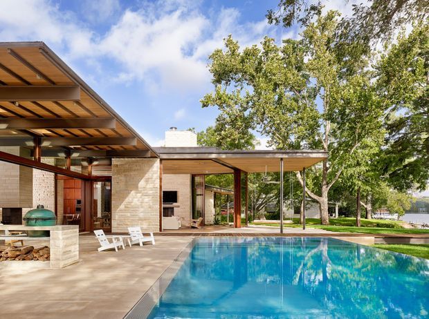 A mid-century-inspired single-storey residence opening onto a large rectangular swimming pool. The image shows broad low-pitched roof overhangs, exposed timber soffits, warm timber and slender dark structural framing, pale masonry walls, extensive glazing, and an outdoor entertaining area with a built-in barbecue and fireplace-like masonry element. Mature trees and a glimpse of water appear beyond the property.