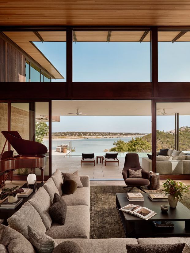 A refined living room with a large sectional sofa, dark wood-framed floor-to-ceiling glazing, and a grand piano. The room opens onto a broad terrace and swimming pool overlooking a coastal bay. Warm timber ceiling slats, dark timber framing, neutral upholstery, a sculptural lounge chair, and a low black coffee table create a luxurious, calm interior with strong indoor-outdoor connection.