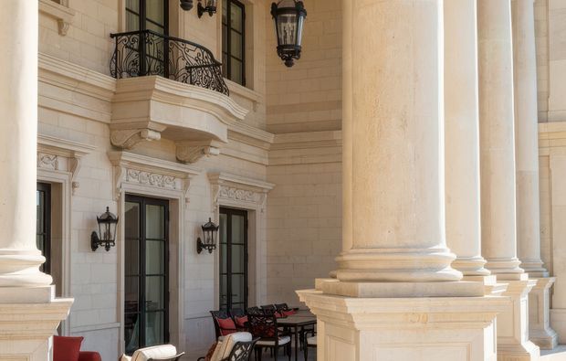 Grand classical exterior portico or terrace featuring tall fluted stone columns with elaborate bases and capitals, a pale limestone block facade, ornate carved window surrounds, French doors, wrought-iron balcony railings, and traditional black wall lanterns. Outdoor dining furniture and seating occupy the covered terrace. The image is strongly characterized by formal, opulent historic architectural detailing.