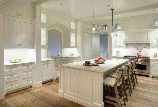 A bright, spacious traditional-style kitchen with extensive cream-painted Shaker cabinetry, a large central island with a pale stone benchtop, and seating for several people. The room includes a freestanding stainless-steel range cooker, integrated storage, pendant lights, timber flooring, a secondary sink area, and an arched architectural opening with display shelving. The overall scheme is warm, refined, and highly functional.