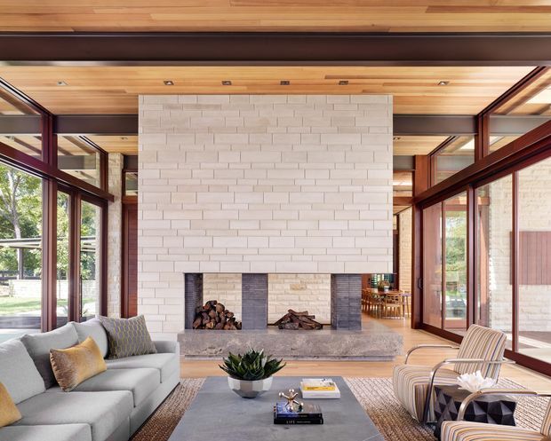 Bright, spacious living room with extensive floor-to-ceiling glazing, warm timber-lined ceiling, dark timber window and door frames, and a large pale brick fireplace wall. A grey sectional sofa, striped lounge chairs, coffee table, and fireplace seating create a refined mid-century-inspired interior with strong indoor-outdoor connection.
