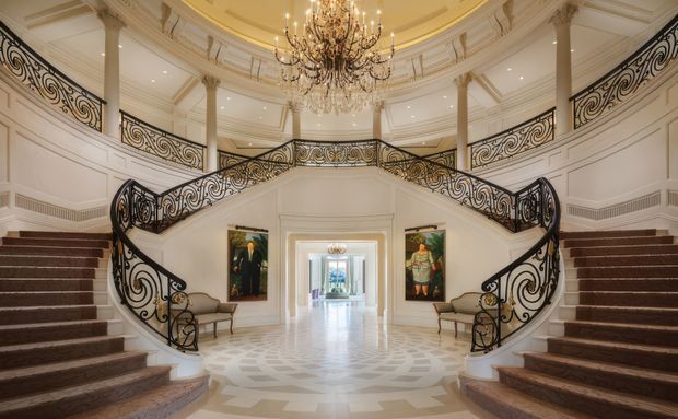 Grand, symmetrical neoclassical-style entry hall with a sweeping bifurcated staircase, ornate black wrought-iron balustrades, white decorative wall panelling, classical columns, patterned pale stone flooring, and a large crystal chandelier. The spacious double-height landing opens toward a bright corridor, with artwork and small upholstered benches along the walls.