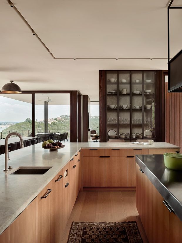 A refined contemporary kitchen with a large U-shaped timber island and cabinetry, pale stone benchtops, an integrated sink and brushed metal tapware. A glazed display cabinet provides storage for crockery, while dark timber wall panelling and black-framed glazing add contrast. The kitchen opens toward a dining area and expansive views beyond.