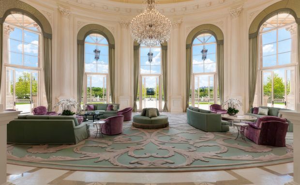Grand, light-filled formal sitting room with a curved bay-like arrangement of tall arched windows and French doors, ornate cream wall panelling, decorative mouldings, a large crystal chandelier, and symmetrical groupings of upholstered seating. A pale patterned carpet, marble or stone perimeter flooring, and muted sage, plum, and cream furnishings create a refined historic-luxury atmosphere. Landscaped grounds are visible through the glazing.