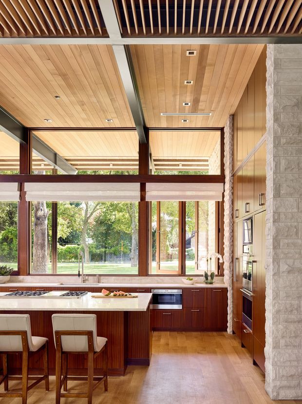 A bright, spacious kitchen with a large central island, integrated cooktop, white stone benchtop, dark timber cabinetry, and timber flooring. Floor-to-ceiling windows and glazed doors open to a leafy garden, while a dramatic timber-lined ceiling with exposed dark structural beams and a slatted timber screen creates strong architectural character. A pale stone-clad wall frames the built-in appliances.