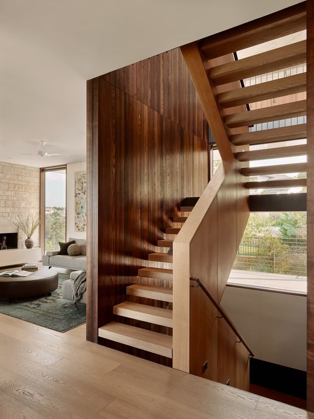 Interior architectural photograph centered on a sculptural timber staircase. Open-riser stairs with a pale timber stringer and handrail rise beside a richly stained vertical timber-panelled wall, with a second flight visible overhead. A bright living room with fireplace, sofa and rug is visible to the left, while large glazing brings in outdoor views on the right. The space has warm natural materials, strong vertical and horizontal timber detailing, and abundant daylight.