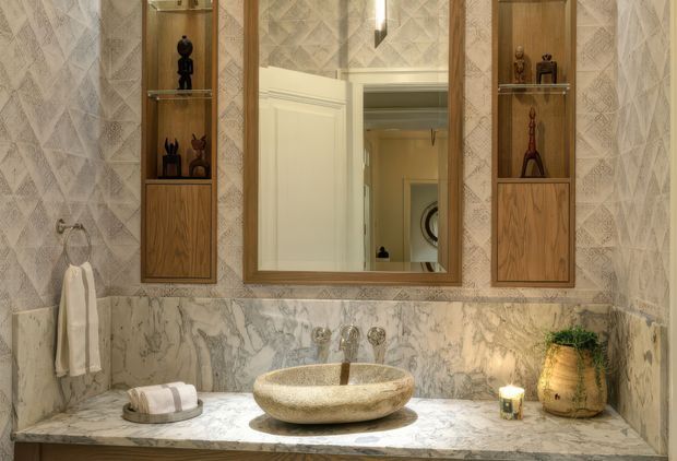A refined bathroom vanity wall featuring a round vessel basin on a marble countertop, a marble backsplash and side walls, a timber-framed rectangular mirror, and symmetrical built-in timber display cabinets with glass shelves. Warm ambient lighting and decorative objects create a luxurious, traditional atmosphere.