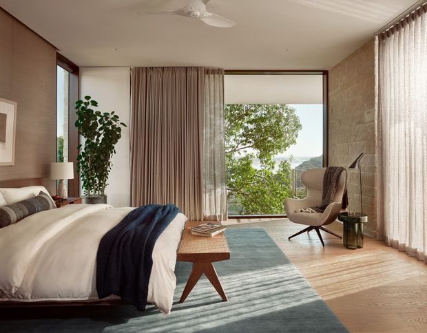 A refined bedroom with a large upholstered bed, layered neutral bedding, a dark blue throw, timber bedside furniture, a sculptural lounge chair, floor-to-ceiling curtains, and expansive corner glazing. A textured light stone wall contrasts with warm timber flooring, while a leafy landscape and distant water or hills are visible outside. The room has a calm, warm modern character with abundant natural light.