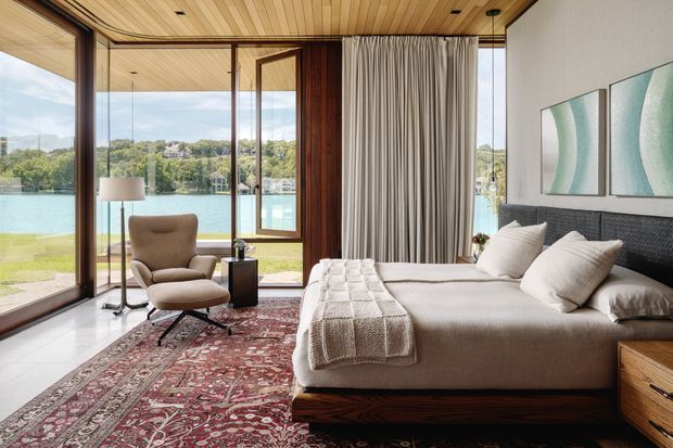 A refined bedroom with floor-to-ceiling glazing overlooking a calm body of water and landscaped surroundings. The room features a low timber bed with upholstered headboard, layered neutral bedding, a patterned Persian-style rug, a sculptural lounge chair, full-height curtains, timber-lined ceiling, and dark timber window and door framing. The composition is warm, serene, and strongly connected to the waterfront view.
