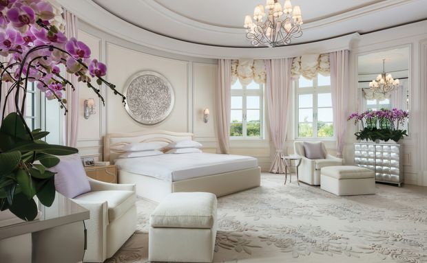 Elegant, bright bedroom with a curved bay of tall windows, pale upholstered bed and seating, ornate white wall panelling, layered blush-pink curtains, floral carpet, crystal chandelier, and orchid displays. The room has a refined luxury aesthetic with symmetrical furnishings and abundant natural light.