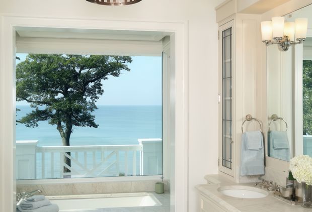 Bright, refined bathroom with a freestanding bathtub positioned beside a large ocean-facing window and white balustrade. The room features pale stone surfaces, white painted cabinetry and wall panelling, a built-in vanity with an oval basin, mirrored panels, traditional chrome fixtures, and decorative wall sconces. A coastal view is visible through the window.