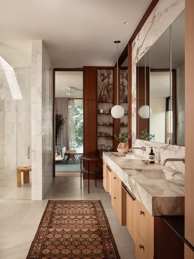 A refined contemporary bathroom with a long double vanity, pale marble-clad walls and benchtop, timber cabinetry, large mirrored panels, and warm dark-wood shelving. Two globe pendant lights hang over the vanity. A glass shower enclosure is visible on the left, while an open doorway leads toward a furnished bedroom or sitting area. The space has a warm, luxurious, hotel-like atmosphere with a patterned runner rug.