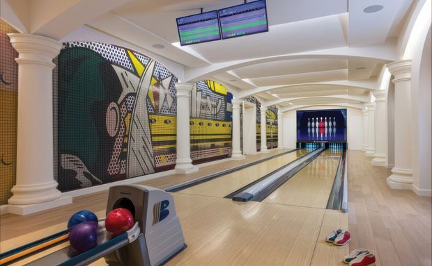 A polished indoor bowling alley with multiple lanes, overhead scoring monitors, ball return equipment, bowling balls and pins. The space has a bright, upscale fit-out with classical fluted columns and arches, decorative mural walls, and light timber-toned lane flooring.