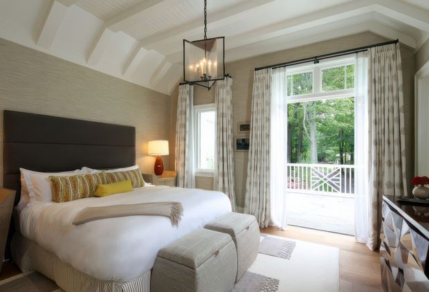 A refined bedroom with a large upholstered dark headboard, white bedding, patterned accent cushions, and a pair of upholstered ottomans at the foot of the bed. The room features a vaulted, beam-accented ceiling, textured neutral wall finish, pale timber flooring, a hanging glass-and-metal lantern, bedside lighting, patterned full-length curtains, and French doors opening to a covered balcony or terrace with a white railing. The overall look is warm, elegant, and traditional with contemporary furnishings.