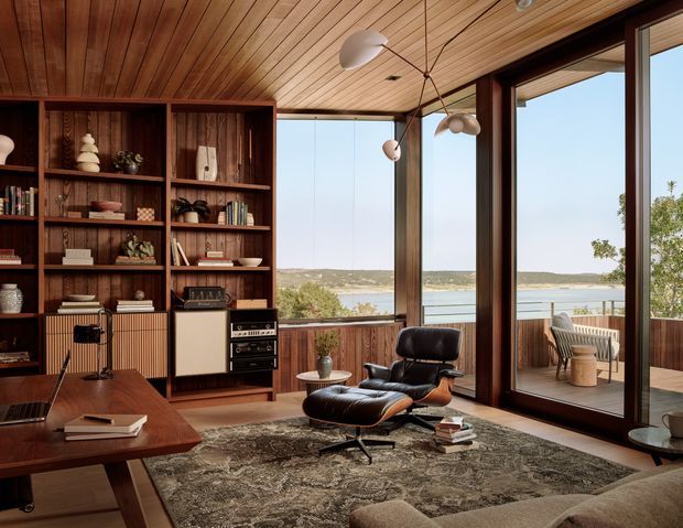A warm mid-century-inspired living room with extensive dark timber built-in shelving, a timber-lined ceiling, vintage audio equipment, and a leather lounge chair with ottoman. Full-height glazing opens to a balcony overlooking a calm waterfront landscape. A large patterned rug, low timber coffee table, and sculptural pendant lighting complete the refined interior.