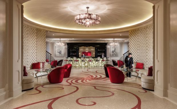 A luxurious, symmetrical hotel or hospitality lounge with a broad open seating area, curved architectural walls, ornate crystal chandeliers, illuminated cove ceiling, patterned carpet with sweeping red motifs, and sculptural red lounge chairs. A bar or reception counter with bottles and glassware is visible at the rear, framed by dark cabinetry and decorative artwork.