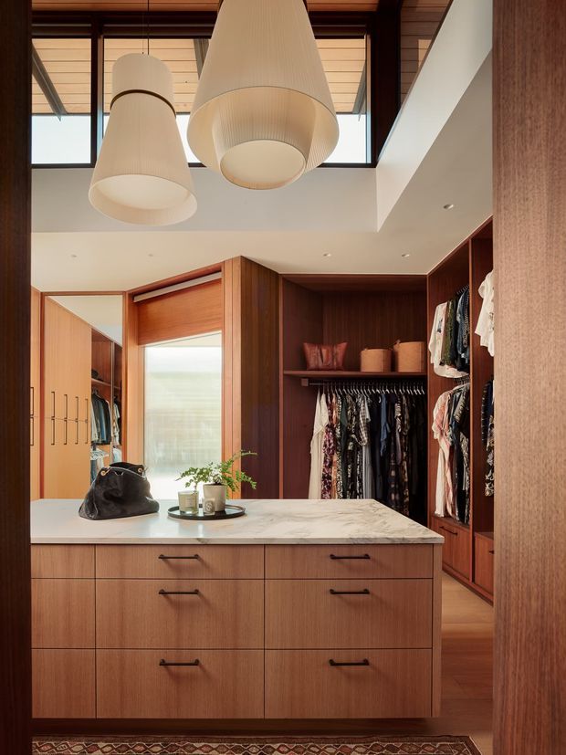 A refined walk-in wardrobe with extensive dark timber built-in cabinetry, hanging rails, open shelving and a central island of drawers topped with a pale veined stone surface. Clothing and accessories are neatly displayed. Large pendant lights hang from a warm timber-lined ceiling, with clerestory glazing and a double-height opening bringing in natural light.