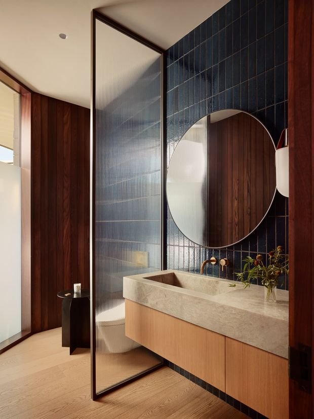A refined contemporary bathroom with a floating timber vanity, thick light stone benchtop and integrated basin, brass tapware, and a large round mirror. The bathroom is enclosed by dark blue vertically set tiles and rich dark timber wall panelling, with a fluted translucent glass screen separating the vanity from the adjacent area. Light timber flooring and warm natural light create a sophisticated, warmly contrasting interior.