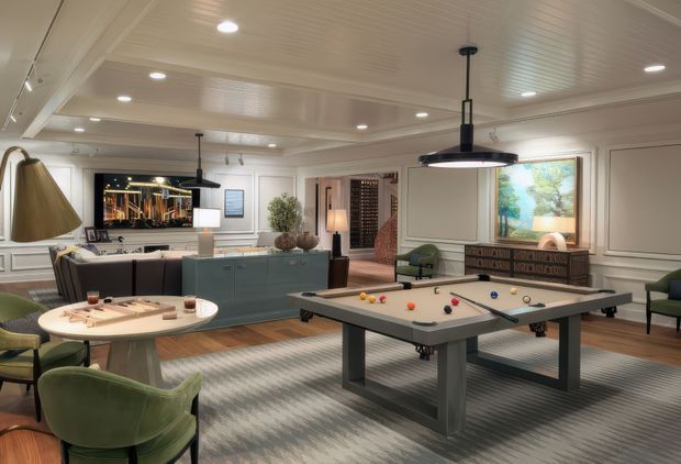 A refined entertainment room with a full-size pool table as the central feature, surrounded by green lounge chairs, a round game table, a bar or kitchenette with blue-grey cabinetry, a large television, decorative lighting, and framed artwork. The room has white panelled walls and a coffered-style panelled ceiling with recessed lighting, creating a polished, club-like atmosphere.
