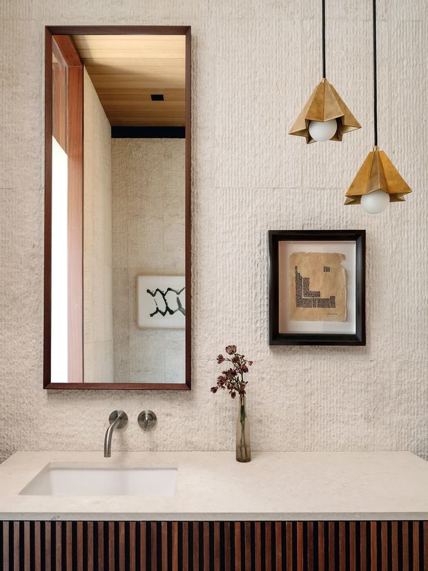 A refined bathroom vanity with a rectangular undermount sink set into a pale stone benchtop. The textured light wall is paired with a warm timber-lined ceiling and dark timber-framed mirror. Two sculptural brass pendant lights hang above the vanity, with framed artwork and a narrow vase adding decorative detail. The vanity front features vertically fluted dark timber.